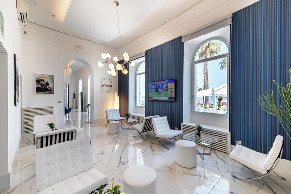 Un vestíbulo de hotel moderno con piso de mármol, mobiliario blanco, un televisor y grandes ventanales con vista a palmeras.