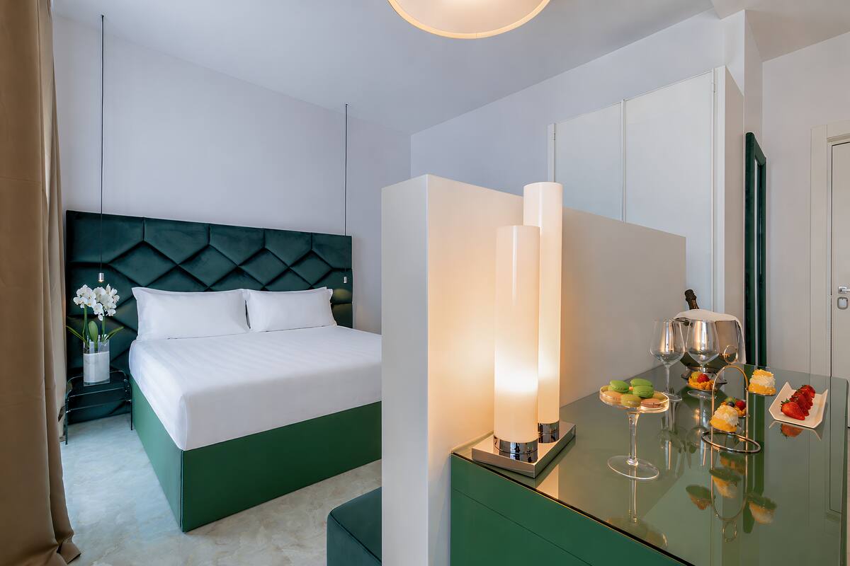 Une chambre d’hôtel avec un lit, une table de chevet avec une lampe, une table en verre avec des verres à vin et des fruits, et une tête de lit verte.