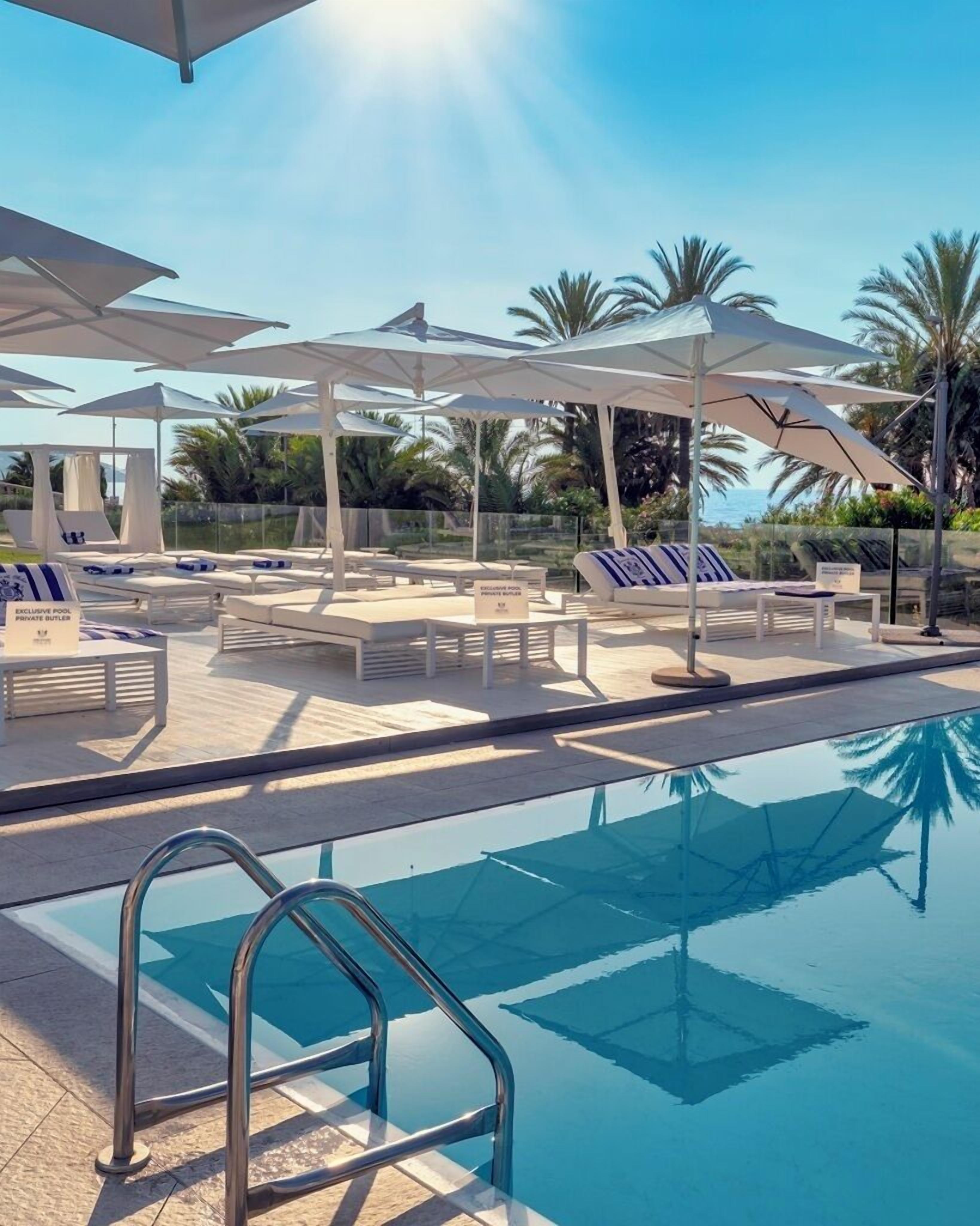 A poolside area with sun loungers and umbrellas, a clear sky, and palm trees in the background.