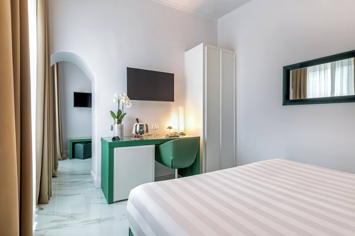 Une chambre d’hôtel équipée d’un lit, d’un bureau, d’une chaise, d’une télévision et d’une petite table sur laquelle est posé un vase de fleurs.