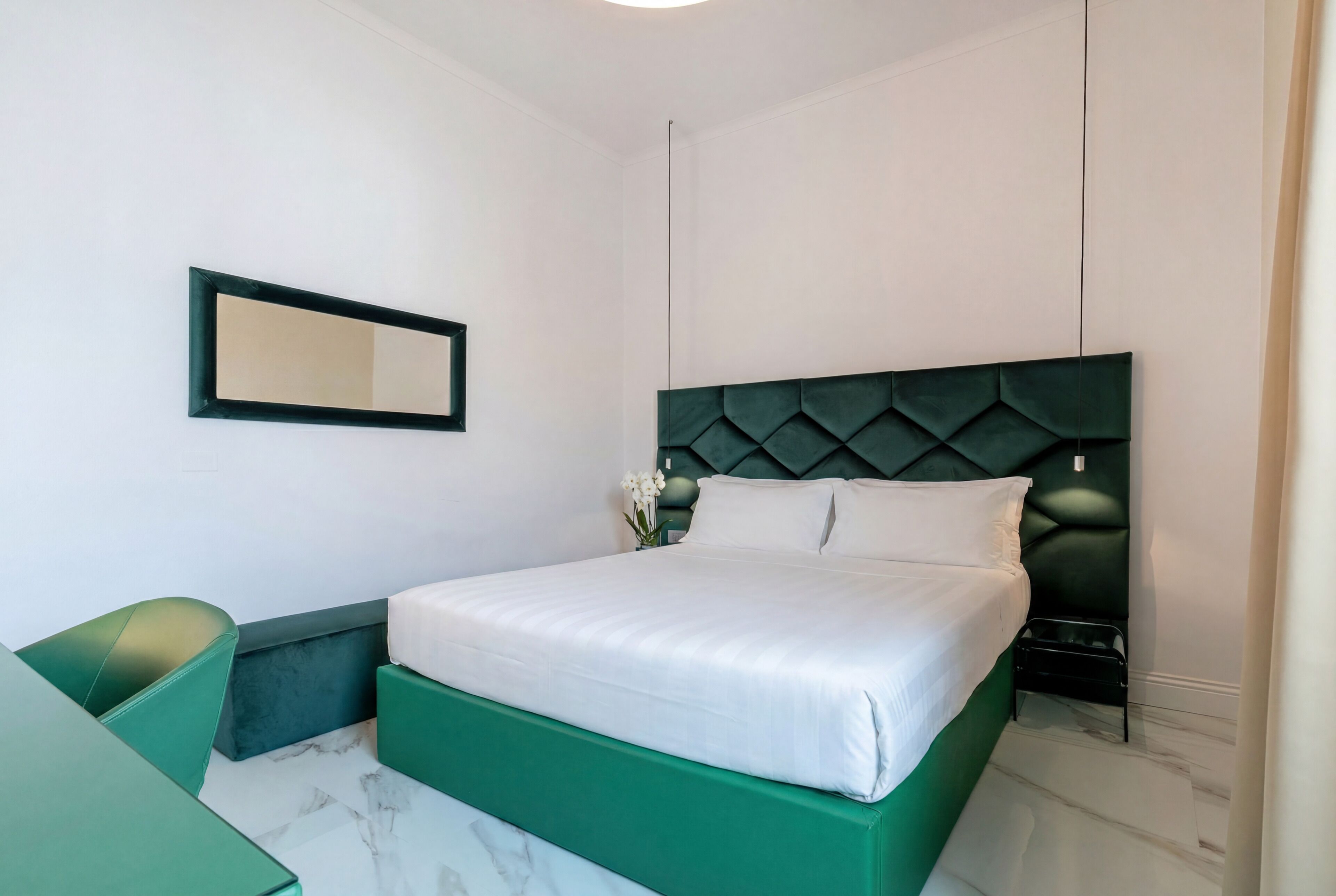 Una habitación con cama, una silla, una mesita, un espejo y un florero con flores.