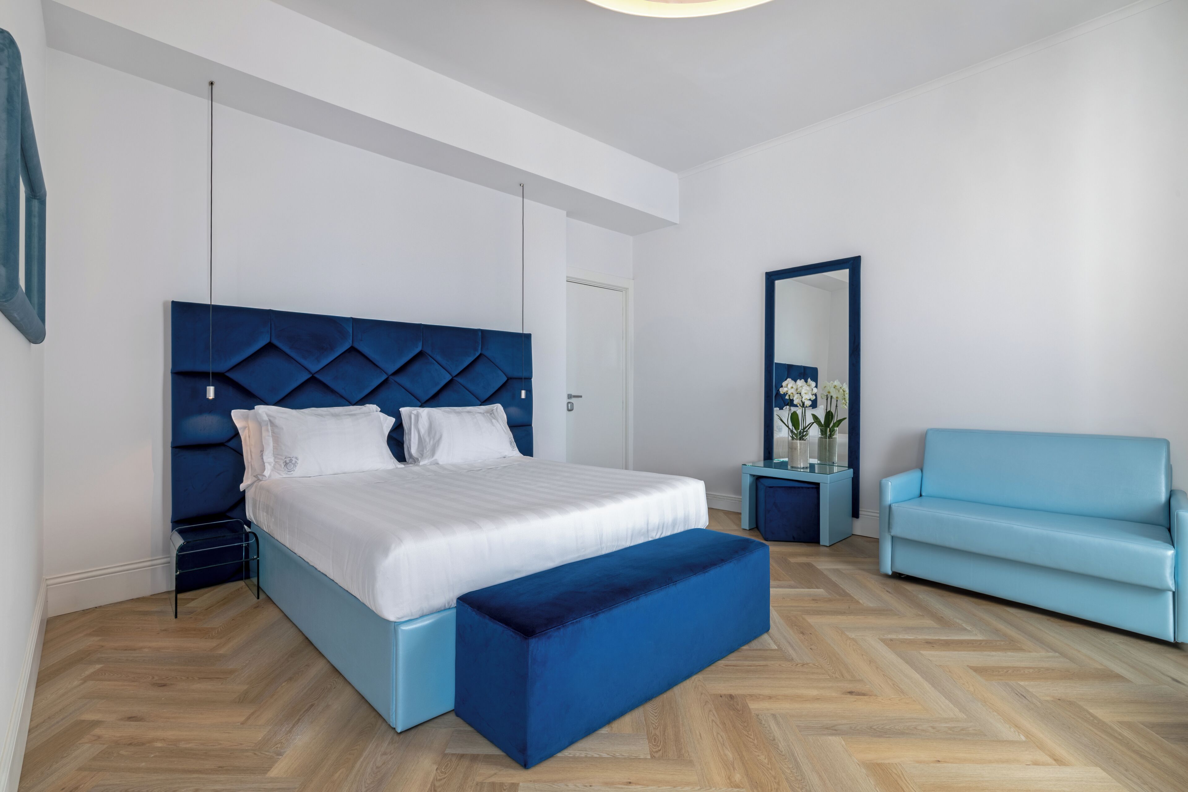 A modern bedroom with a large bed, a blue bench, a sofa, a bedside table, and a mirror.