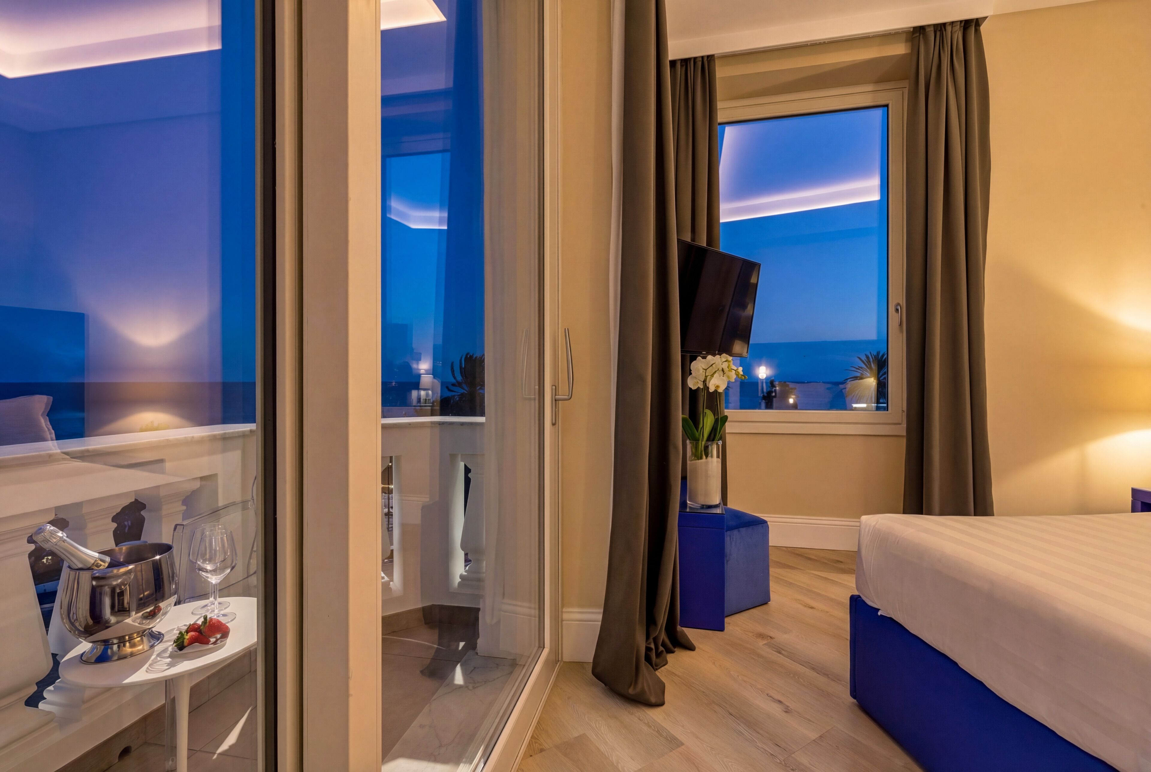 A hotel room with a bed, a small table with a bottle and glass, a TV, and a window with curtains.