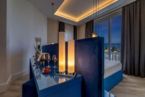 A modern hotel room with a large bed, a glass table with candles, and a view of the sea.