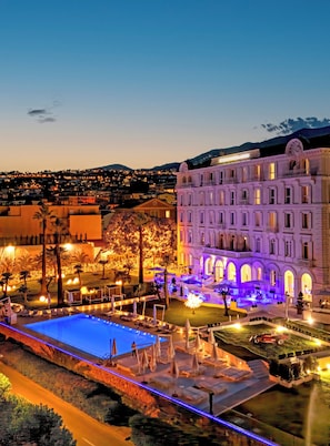 A hotel with a swimming pool, outdoor seating, and a view of the city at dusk.