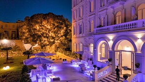 Un espace extérieur bien éclairé, aménagé avec des tables et des chaises, un grand arbre décoré de guirlandes lumineuses et un imposant bâtiment doté d’arches et de balcons.