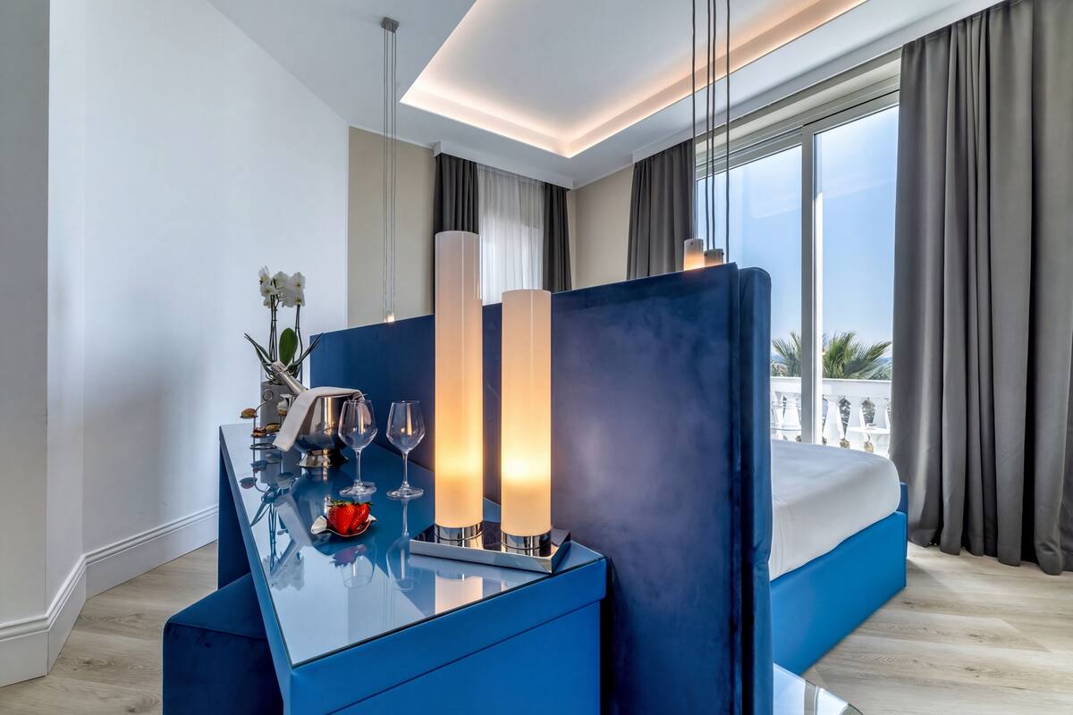 Une chambre d’hôtel moderne avec un grand lit, une console bleue et une vue sur des palmiers.