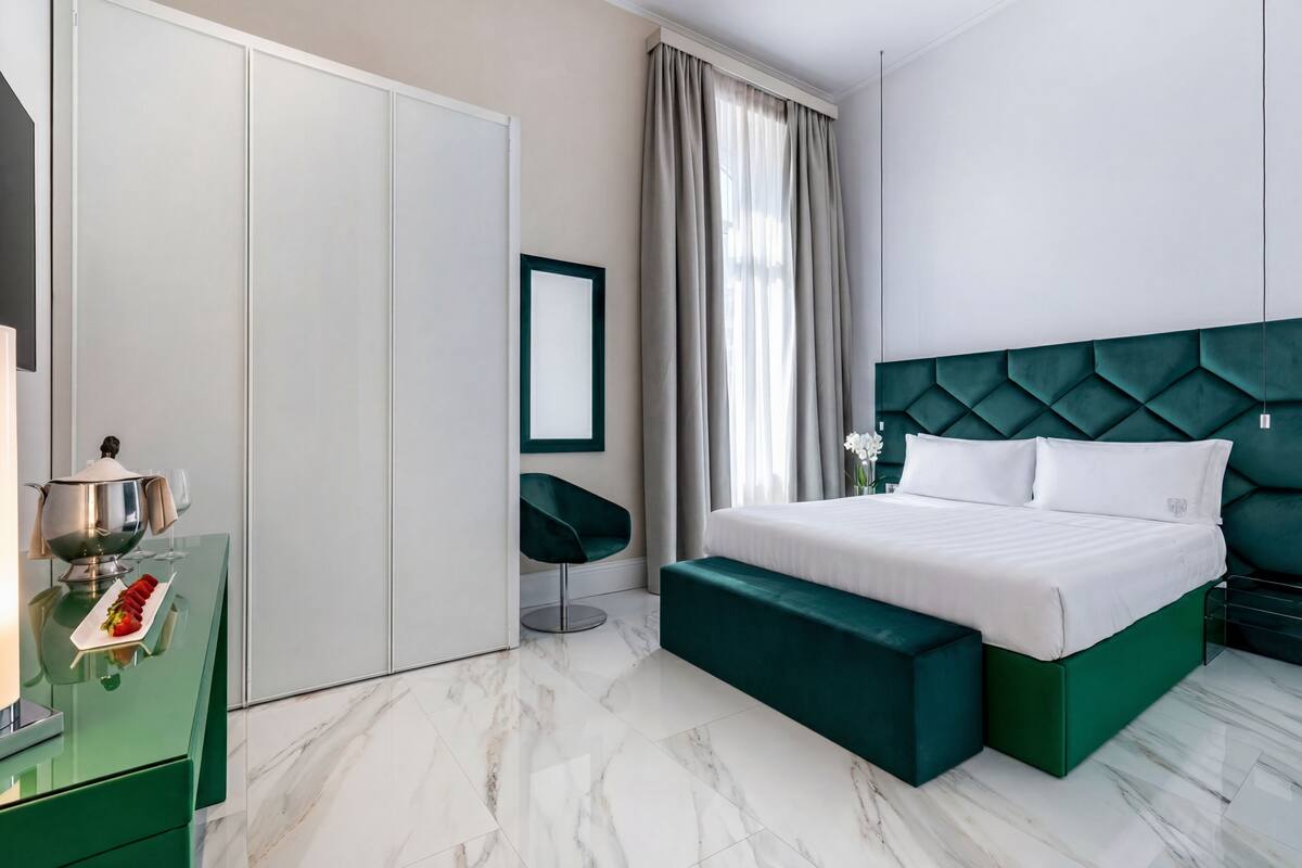 Une chambre moderne avec un lit recouvert d’un tissu vert, une tête de lit blanche, un banc vert, une table de chevet blanche agrémentée d’un pichet et de verres argentés, et une chaise verte.