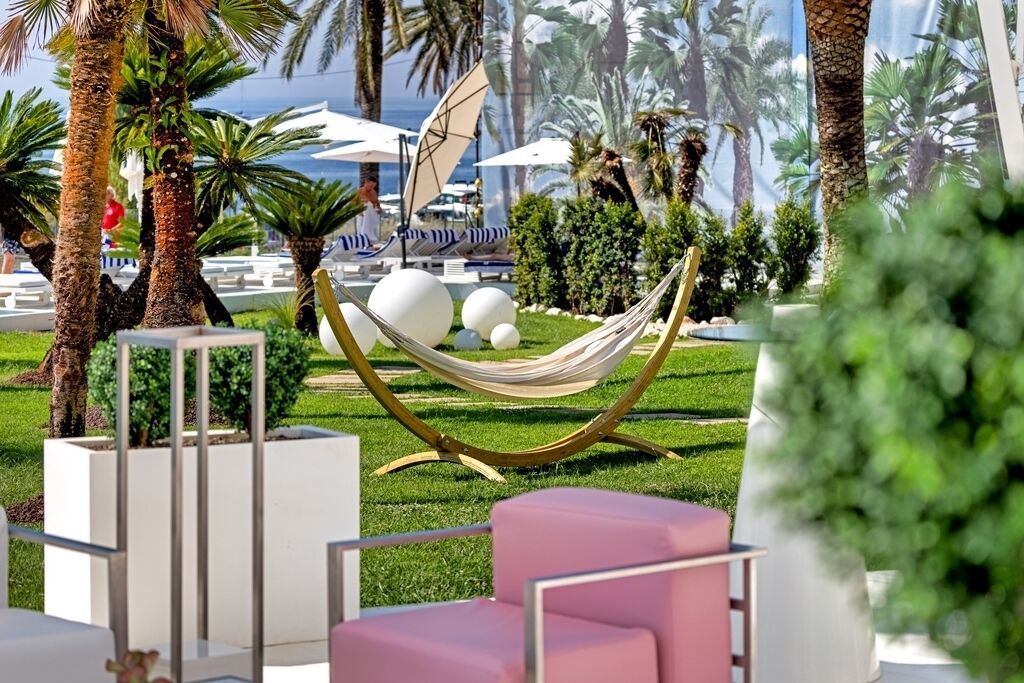 Una hamaca en un jardín exuberante con palmeras, un área de piscina y mobiliario exterior.