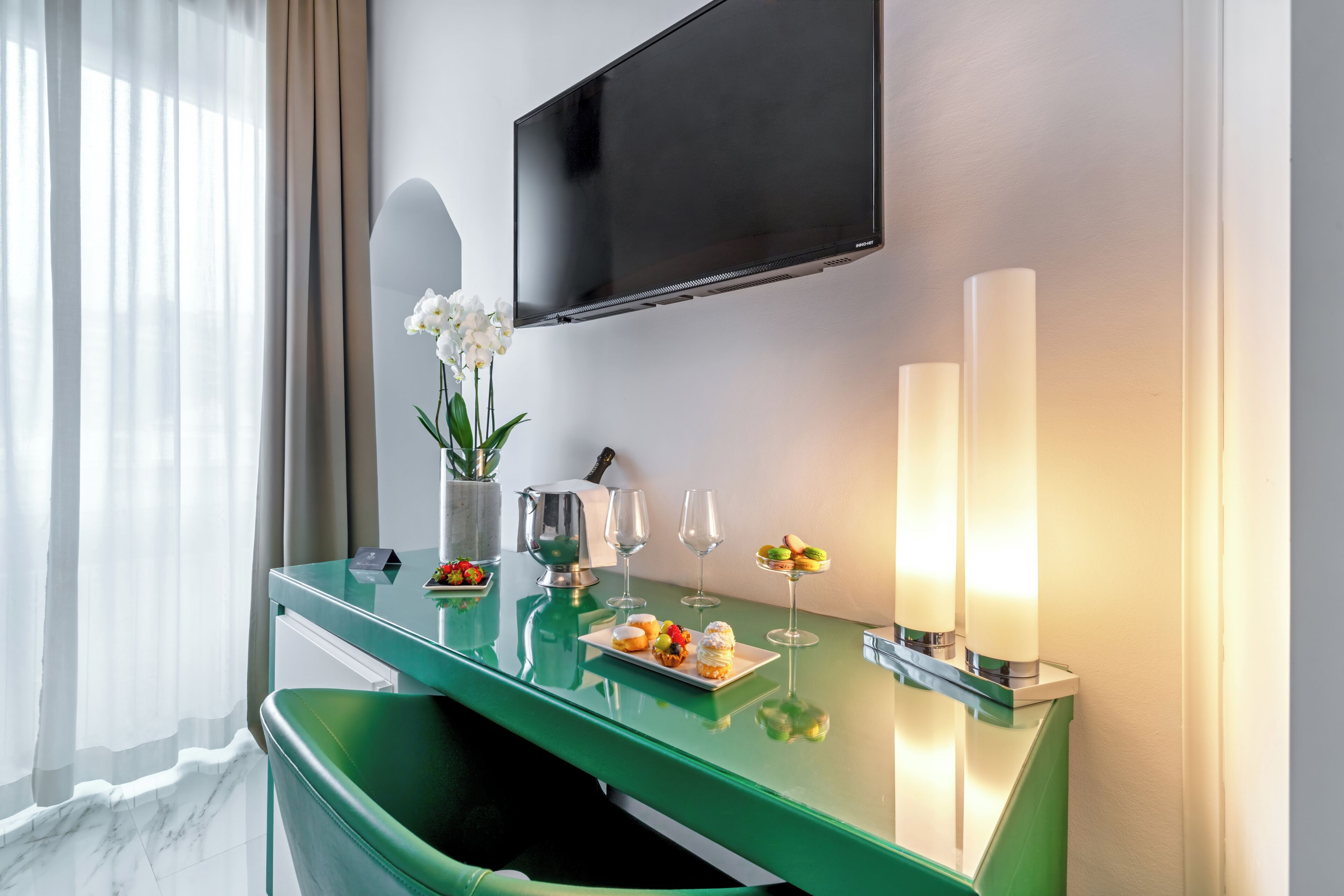 Habitación de hotel moderna con un escritorio verde, un televisor de pantalla plana, un jarrón con flores blancas y una mesa con comida y bebidas.