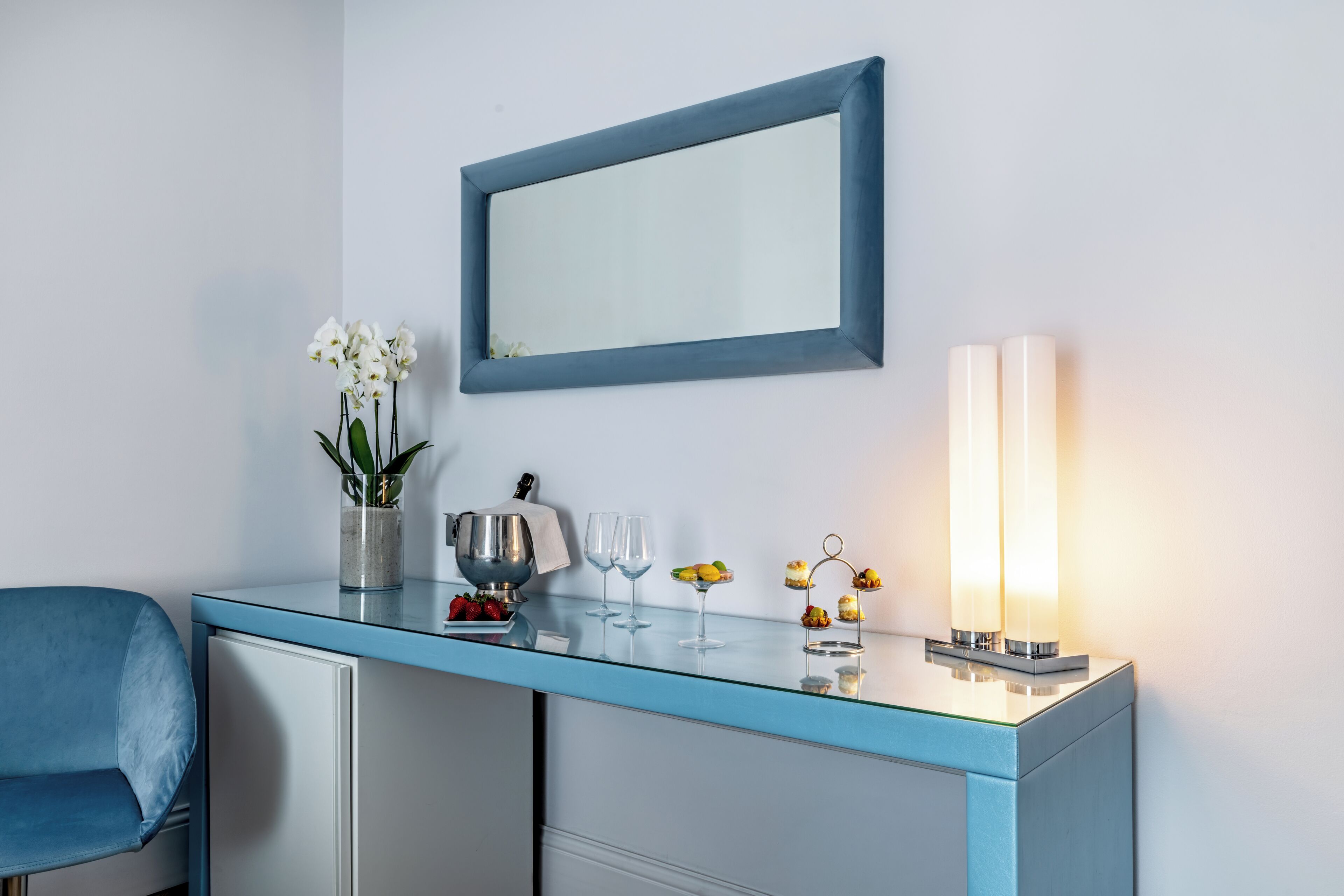 Ein Zimmer mit blauem Glastisch, einem Stuhl, einem Spiegel, einer Vase mit Blumen, einer Schale mit Erdbeeren und einer Kerze.
