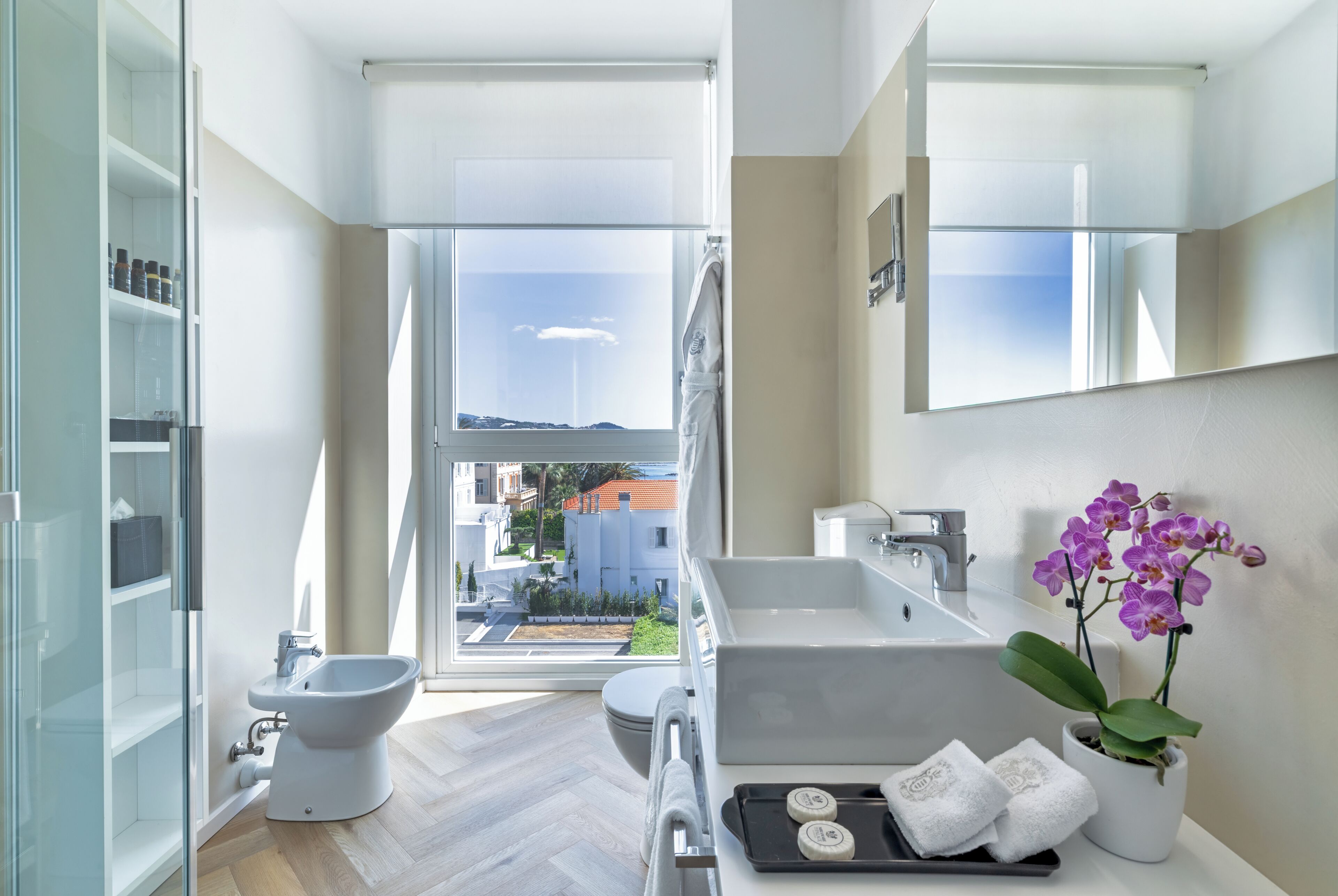 Un baño moderno con bidé, inodoro y bañera, un ventanal con vista a edificios, y una orquídea en maceta.