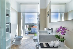 Een moderne badkamer met een bidet, toilet en bad, een groot raam met uitzicht op gebouwen, en een potorkidee.