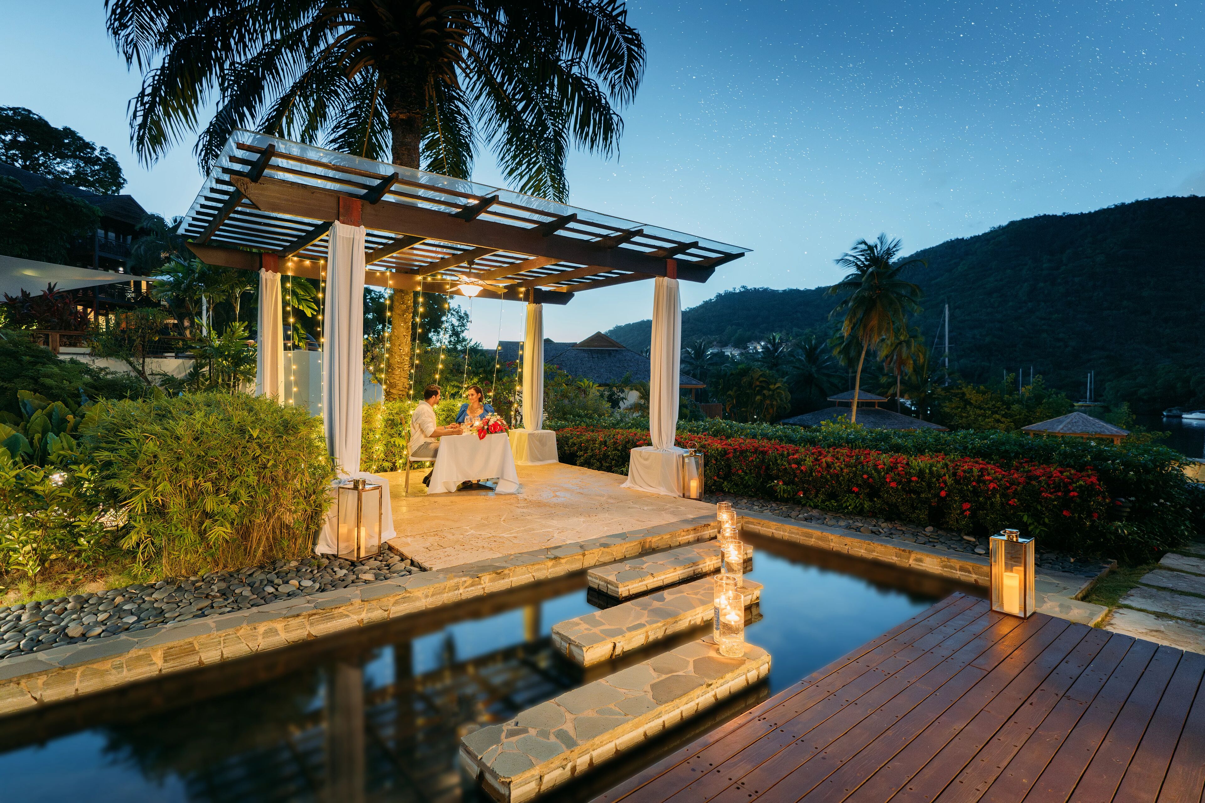 Un pabellón con piscina y terraza de madera, rodeado de vegetación tropical y con montañas al fondo.