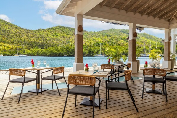 An outdoor dining area with tables and chairs, overlooking a body of water and lush green hills.