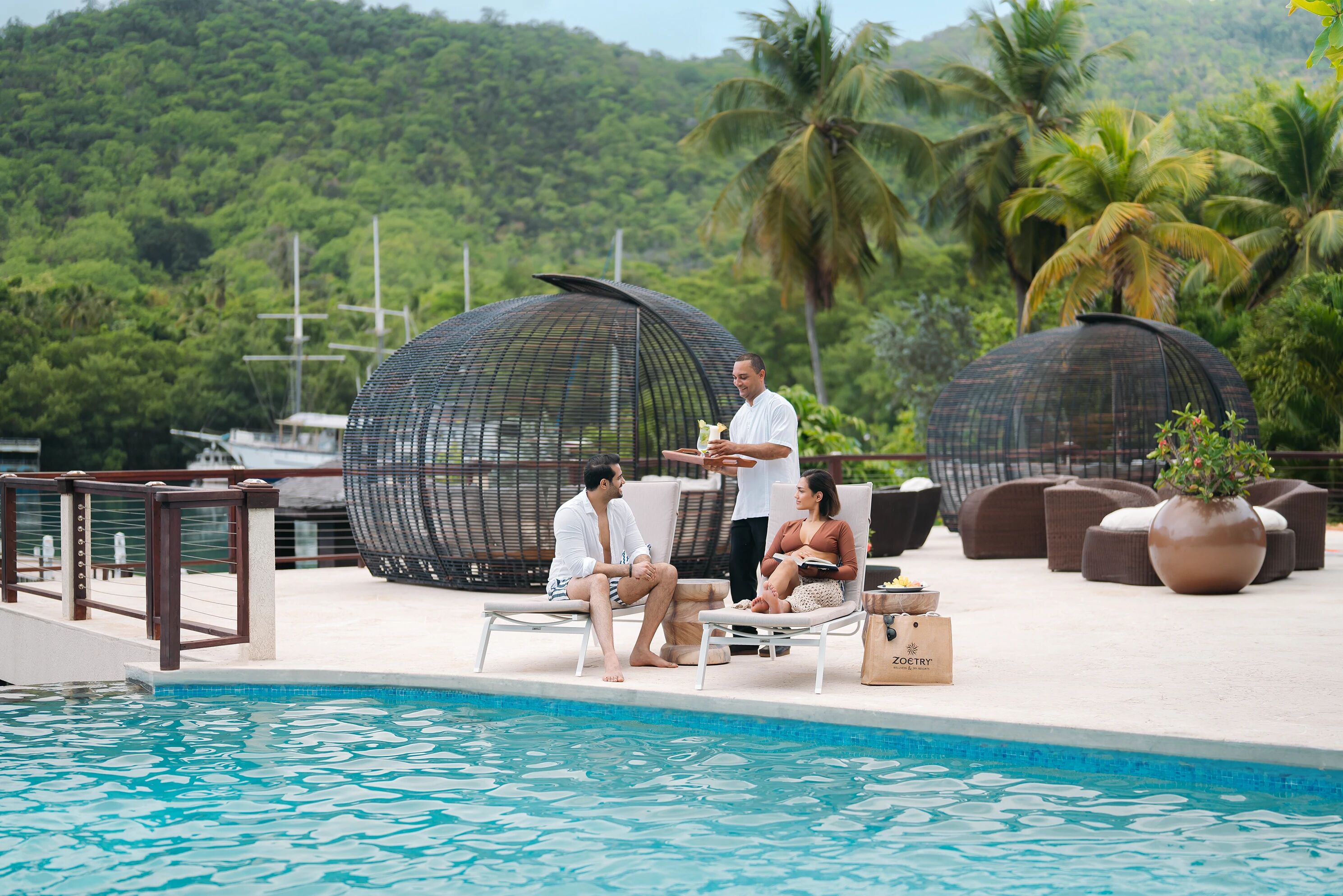 A poolside area with lounge chairs, a waiter serving food, and a view of lush greenery in the background.
