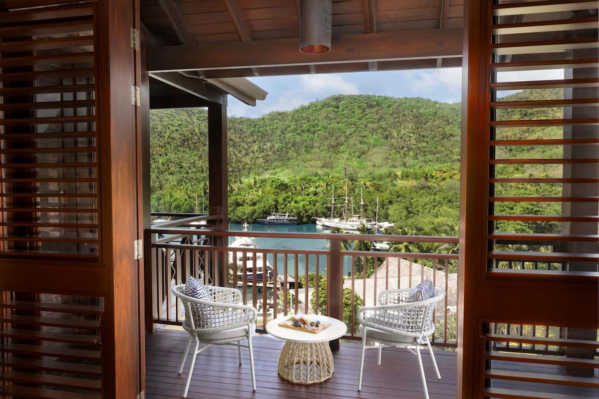 Eine Holterrasse mit zwei weißen Korbstühlen und einem kleinen runden Tisch, mit Blick auf einen Yachthafen mit Booten und einer gebirgigen Landschaft.