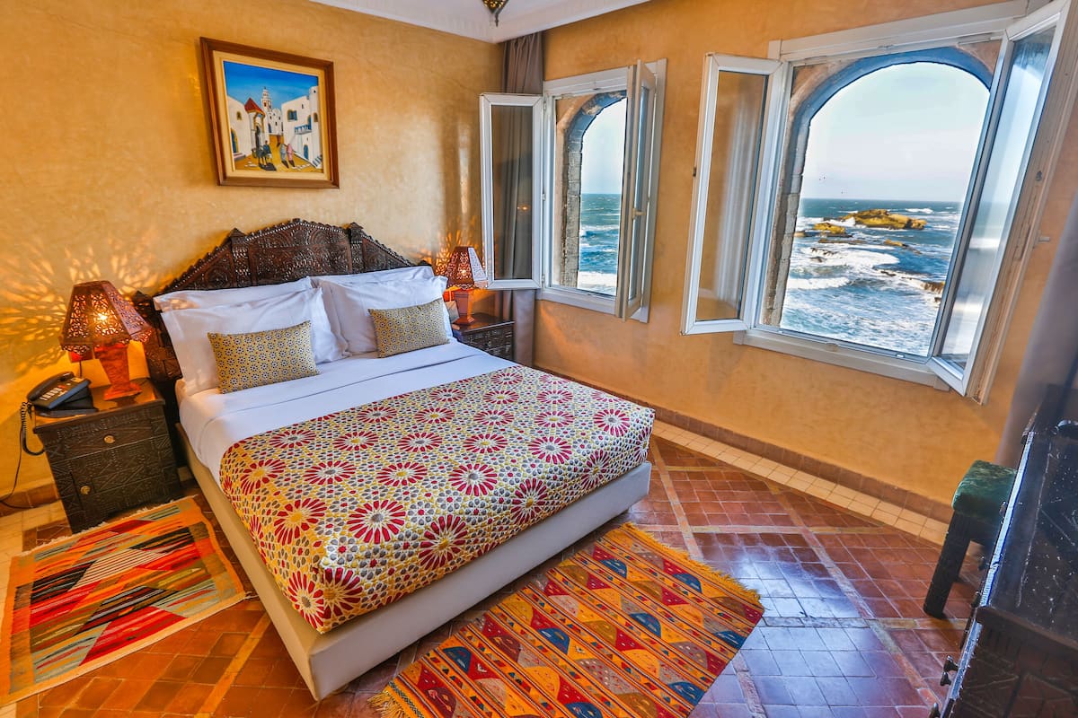 A bedroom with a bed, a nightstand, a lamp, a painting, and a view of the sea through a window.