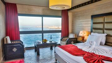 Habitación de hotel con cama, mesita de noche, silla, mesa con lámpara y vista al mar.