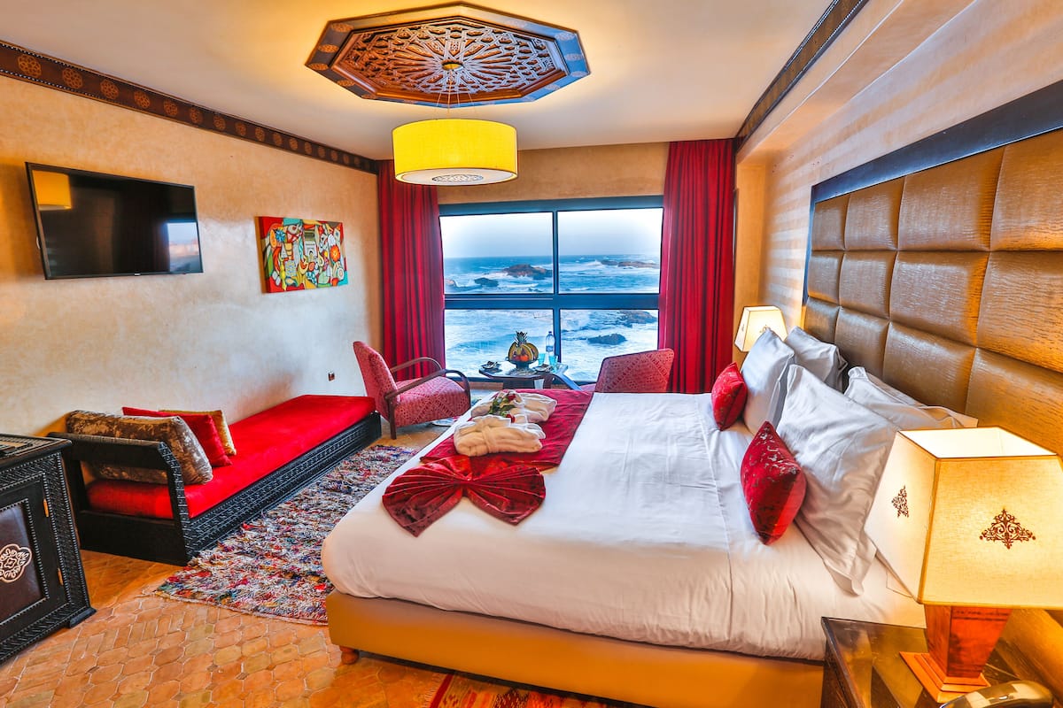 A hotel room with a large bed, a TV, a view of the ocean, and a red sofa.