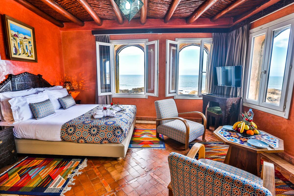 A hotel room with a bed, a chair, a table with fruit, and a view of the sea through open windows.