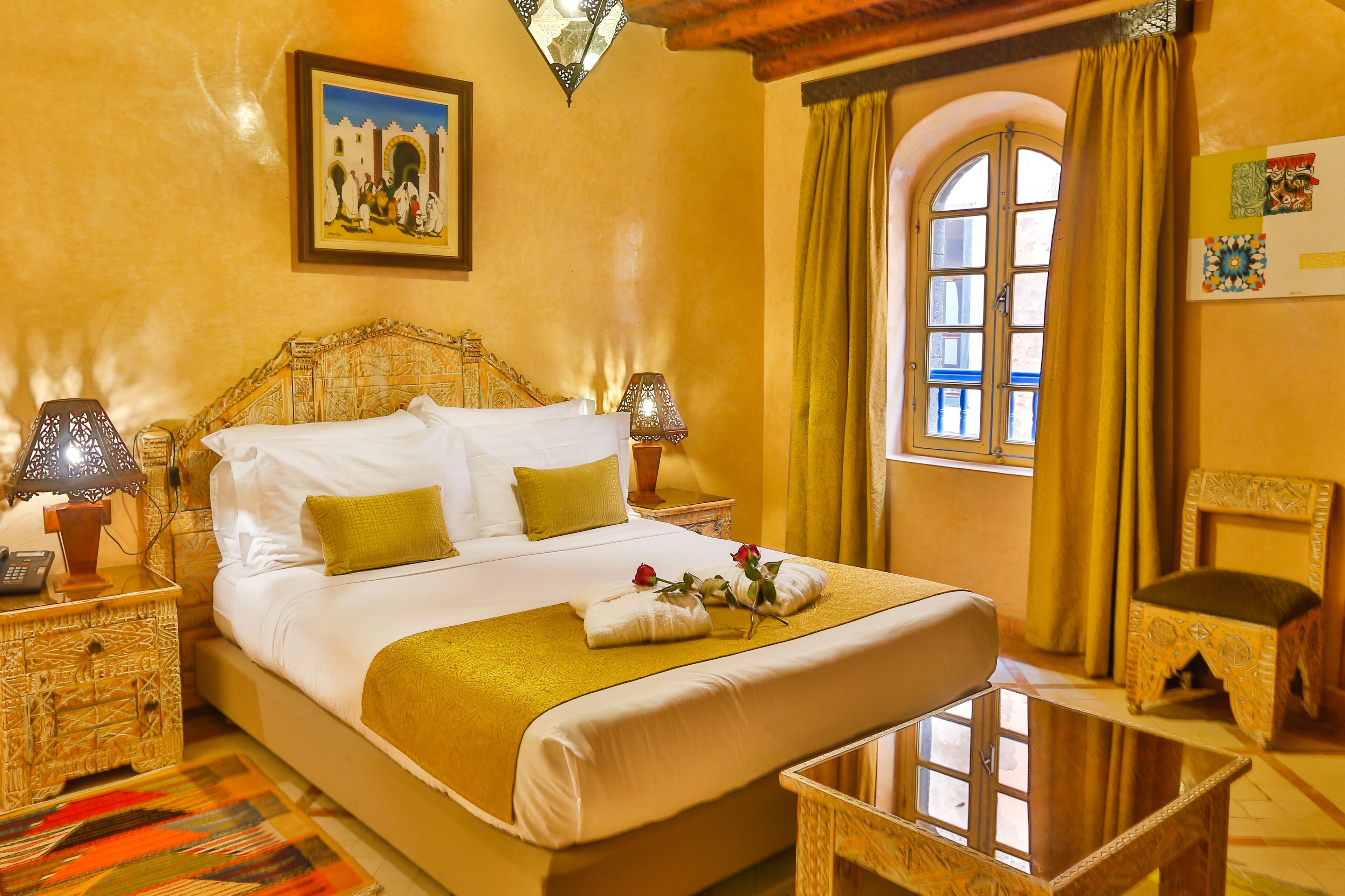 A hotel room with a large bed, two bedside tables with lamps, a small table, a chair, a window with curtains, and a wall-mounted picture.