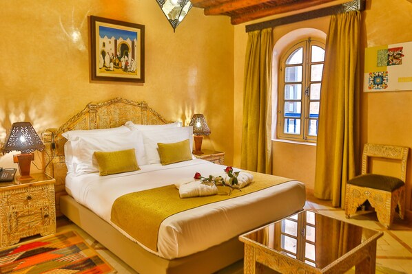 Une chambre d’hôtel avec un grand lit, deux tables de chevet avec des lampes, une petite table, une chaise, une fenêtre avec des rideaux et un tableau accroché au mur.
