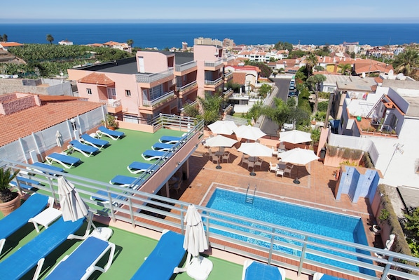 A rooftop pool area with lounge chairs and umbrellas, overlooking a coastal cityscape.