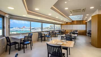 Un espace de restauration moderne, avec des tables et des chaises, de grandes fenêtres donnant sur un port et des navires, et un plafond vitré.