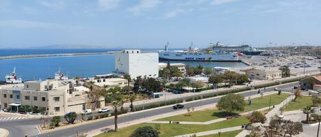 A coastal cityscape with a prominent white building with blue accents, a marina with cruise ships, and a road with cars and pedestrians.