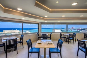 En restaurang med bord och stolar, stora fönster med utsikt över havet och en avlägsen stadsbild.