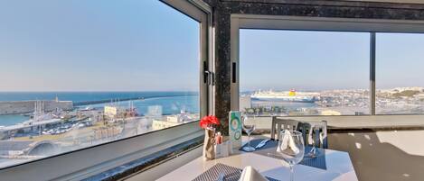 Un comedor con una mesa lista para dos personas, un jarrón con flores rojas y vistas al mar y al paisaje urbano.