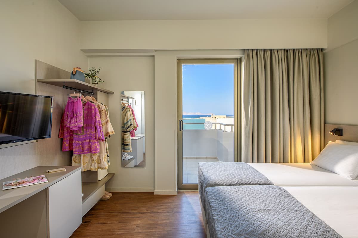 A hotel room with a bed, a TV, a desk, and a balcony with a view of the sea.