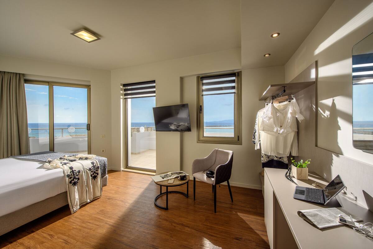 A hotel room with a bed, a desk with a laptop, a chair, a TV on the wall, and a view of the sea through large windows.