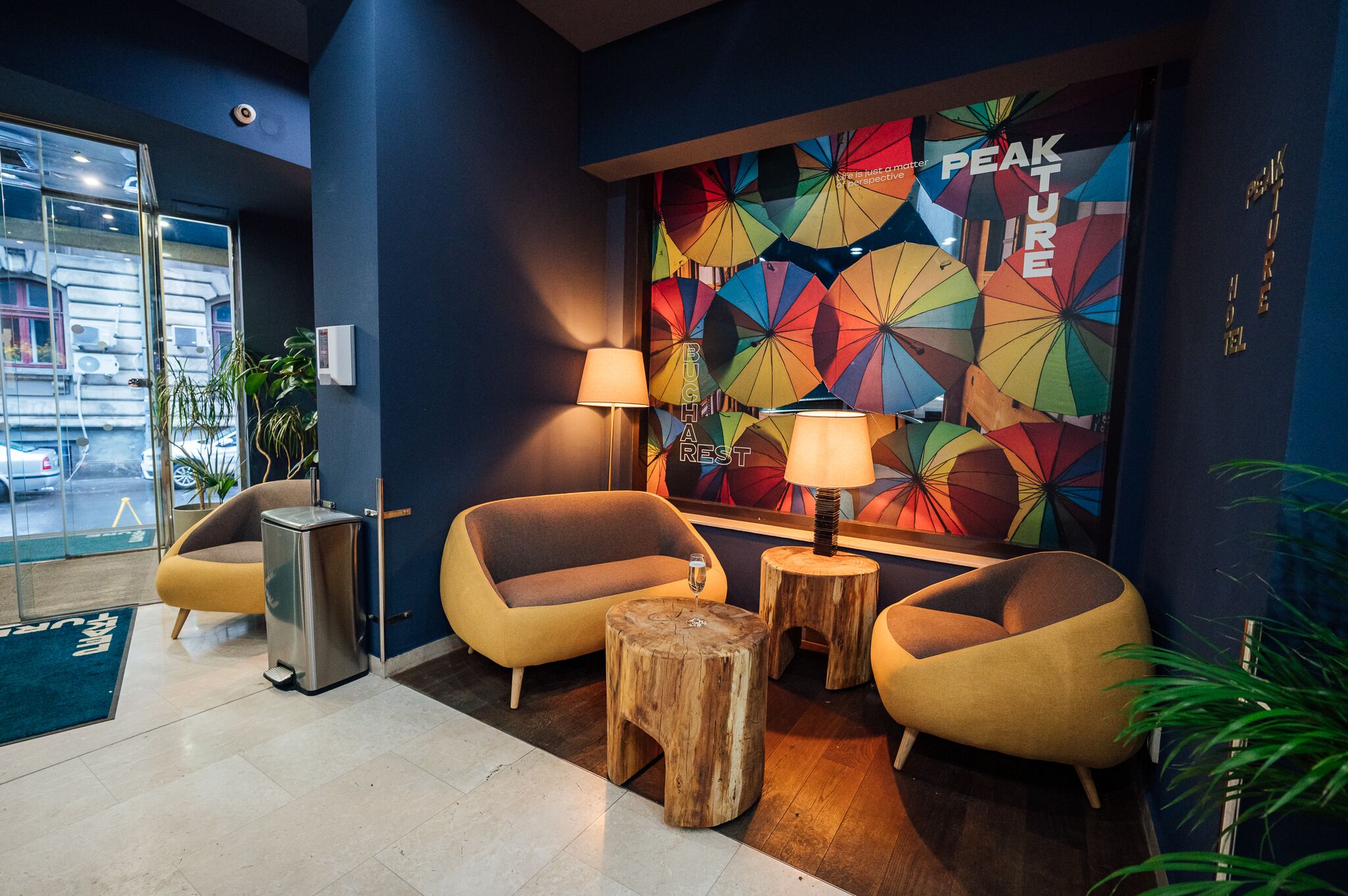 A modern lobby with two yellow armchairs, a wooden stool, and a wall mural with colorful umbrellas.
