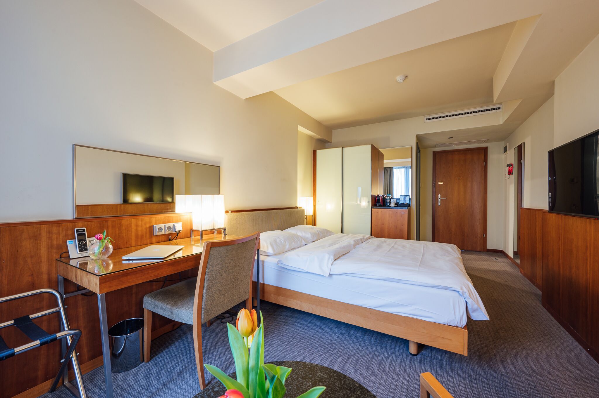 A hotel room with a bed, desk, chair, and a small table with flowers.