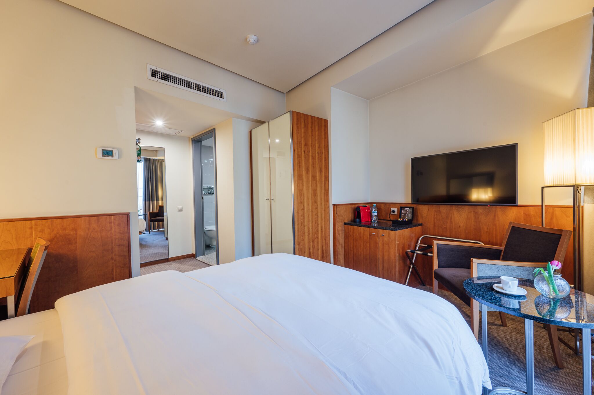 Une chambre d’hôtel avec un grand lit, un bureau avec une chaise, une télévision et une petite table avec une lampe et un vase.
