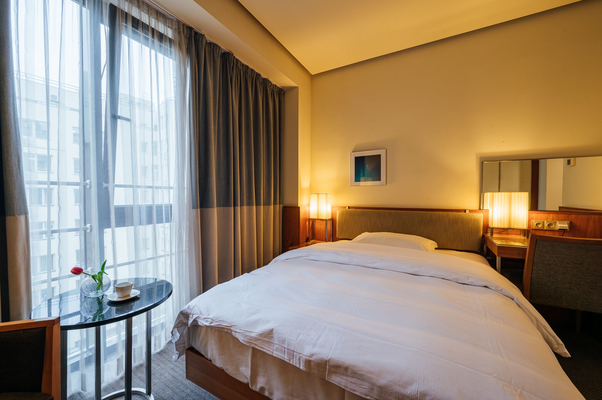 Ein Hotelzimmer mit einem großen Bett, einem kleinen Tisch mit Tasse und Rose, einem Sessel, einem Nachttisch mit Lampe und einem Fenster mit Vorhängen.