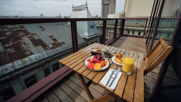 Una mesa de madera con alimentos para el desayuno en un balcón con vistas a un paisaje urbano.
