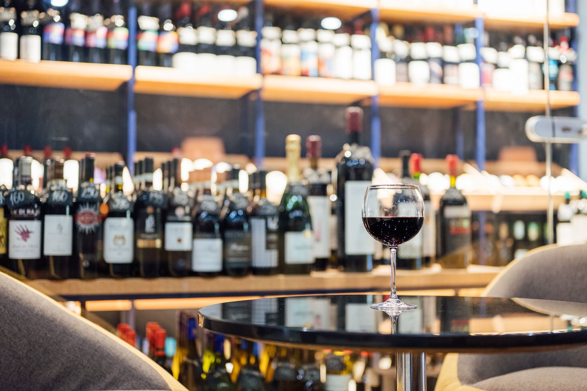 A bar area with a glass of red wine on a table, surrounded by shelves of wine bottles.