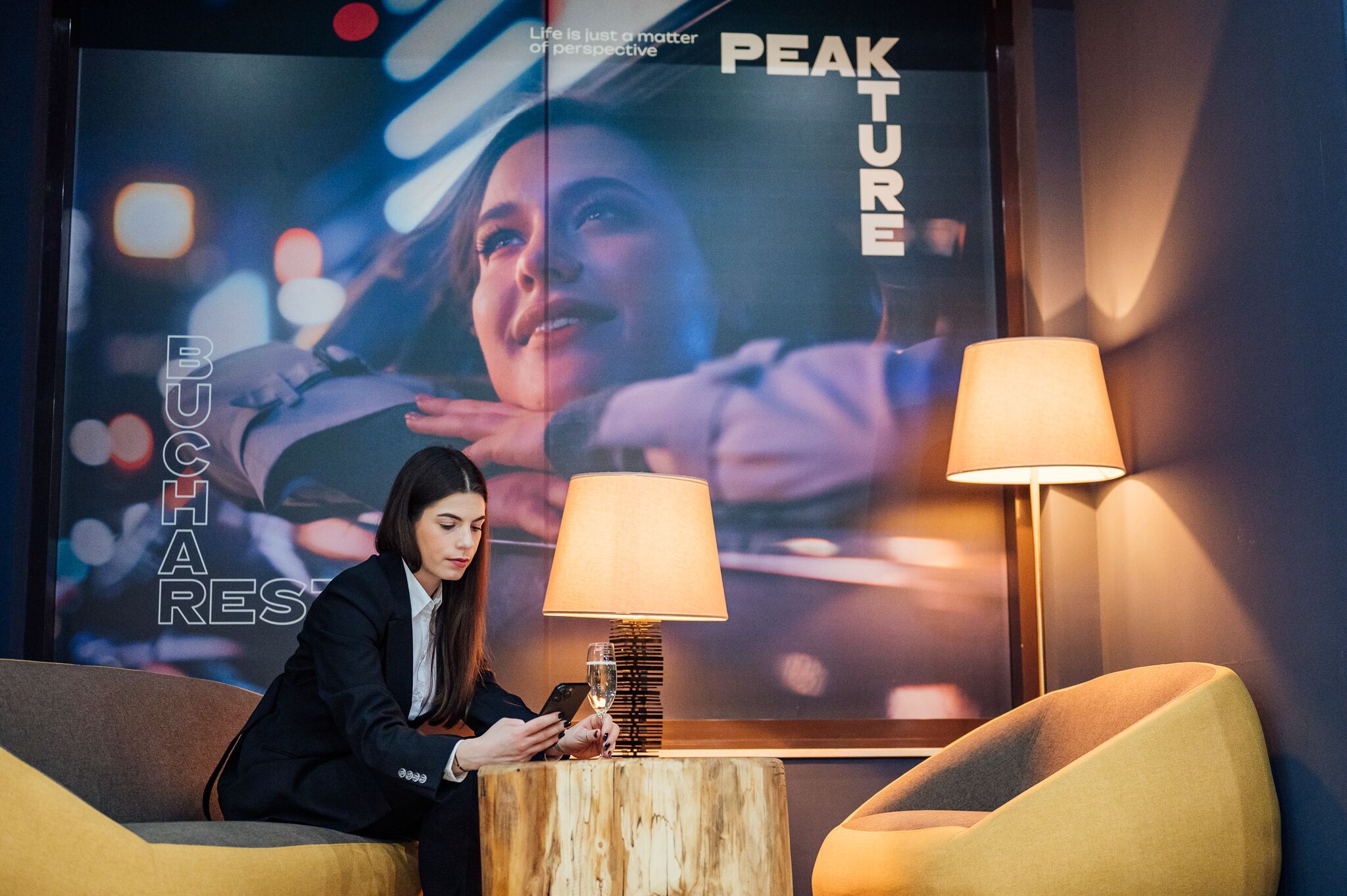 A woman sitting on a sofa in a modern lobby with a large screen displaying 'PEAK TURE' and 'BUCHAREST'. There are two lamps on a wooden table in front of her.