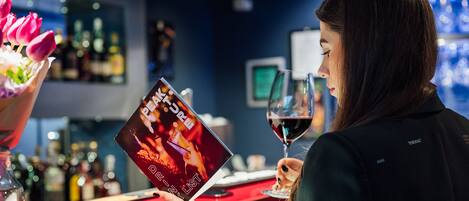 A person holding a menu at a bar with a glass of red wine, surrounded by bottles and a vase of flowers.