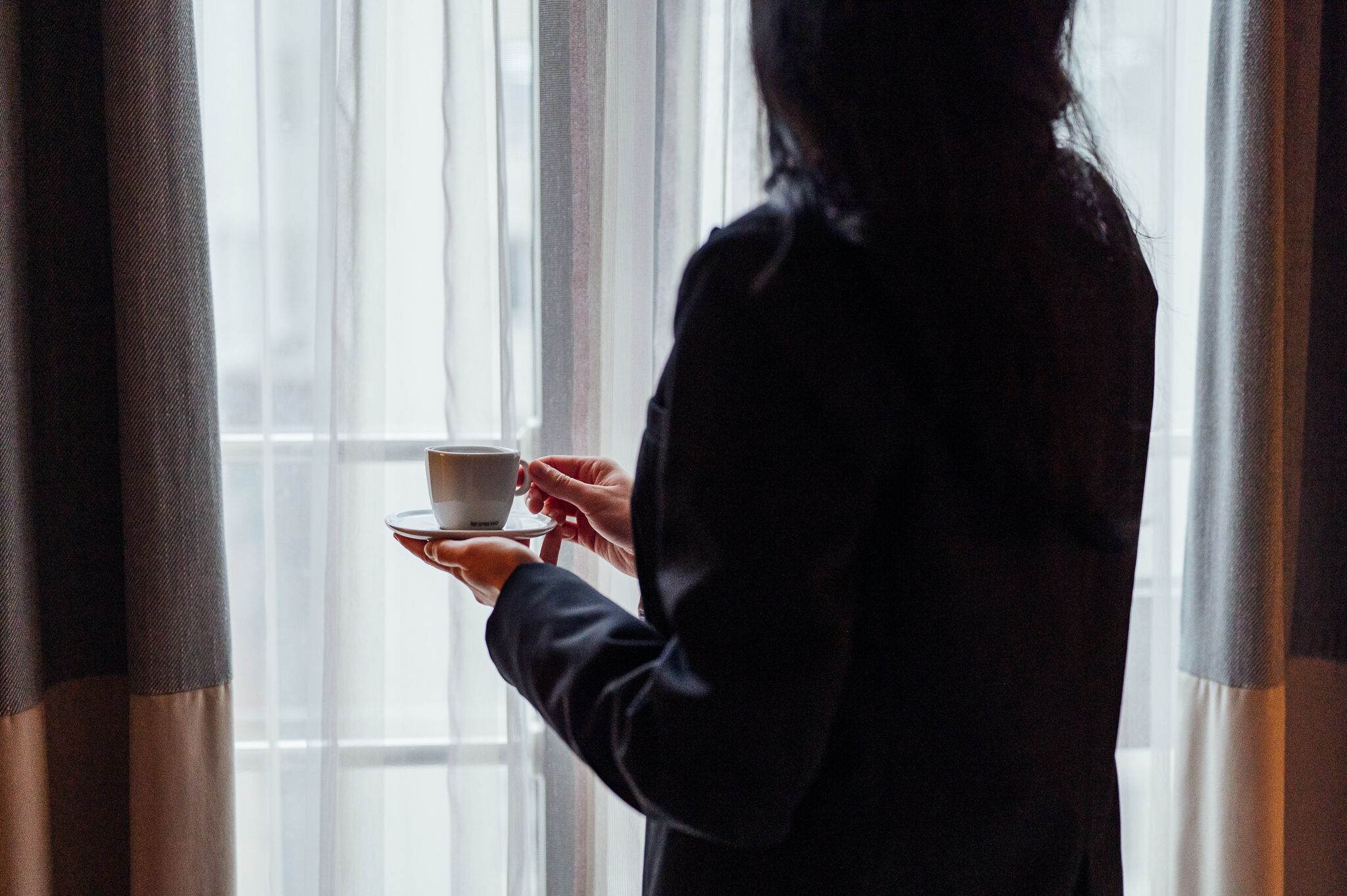 Une personne tenant une tasse près d’une fenêtre avec des rideaux.