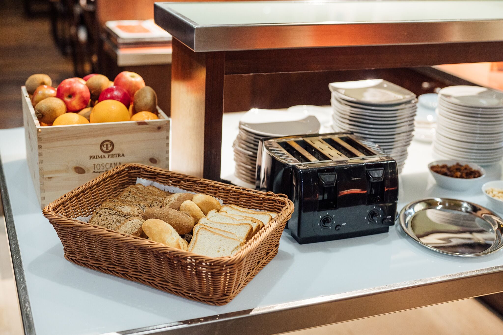 Montaje de bufé con una cesta de mimbre para pan, una caja de frutas, platos apilados y una tostadora.