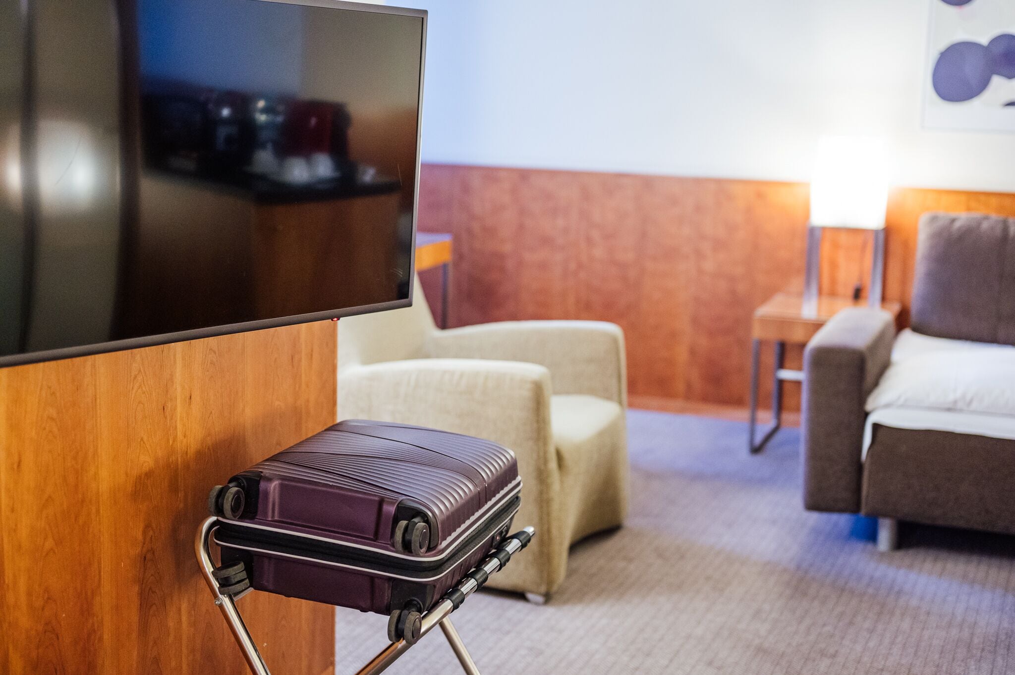 Une chambre d’hôtel avec un téléviseur fixé au mur, un coin salon avec deux fauteuils, une petite table et une valise posée sur un chariot.