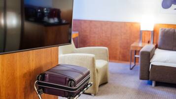Une chambre d’hôtel avec un téléviseur fixé au mur, un coin salon avec deux fauteuils, une petite table et une valise posée sur un chariot.