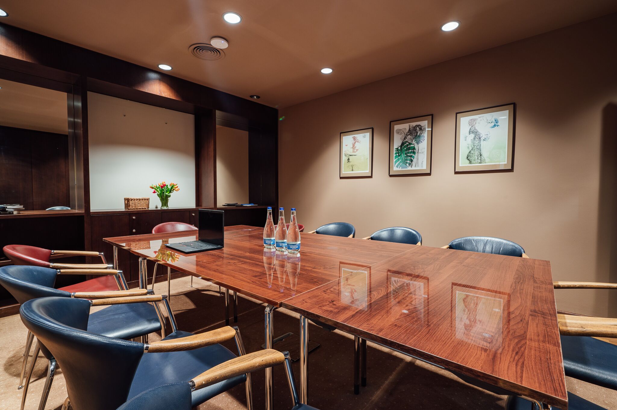 A conference room with a wooden table, blue and red chairs, and framed artwork on the wall.