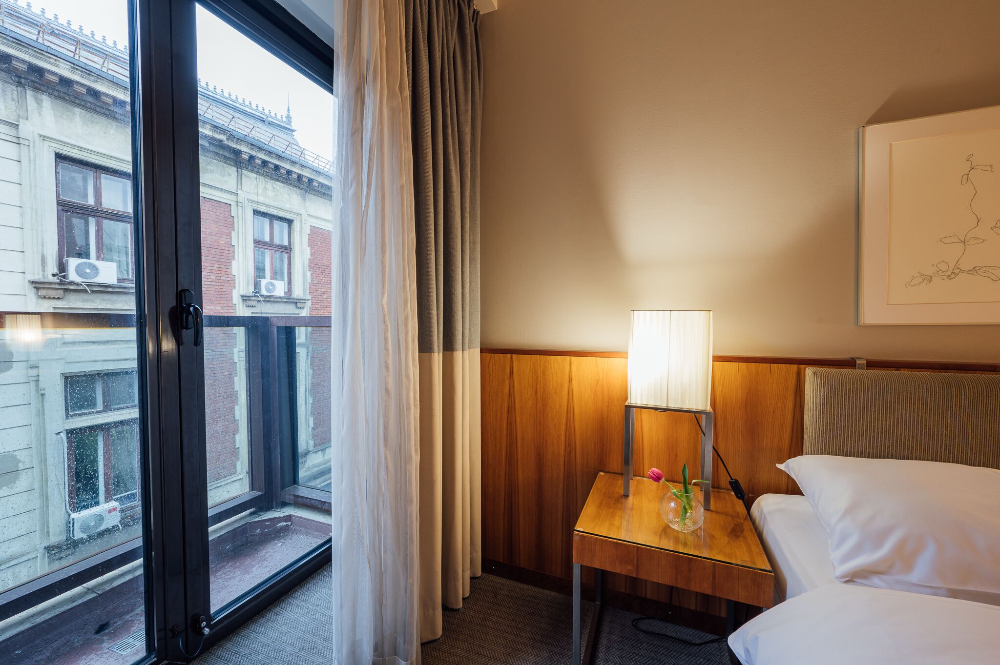 A hotel room with a bed, a bedside table with a lamp, a window with curtains, and a view of buildings outside.