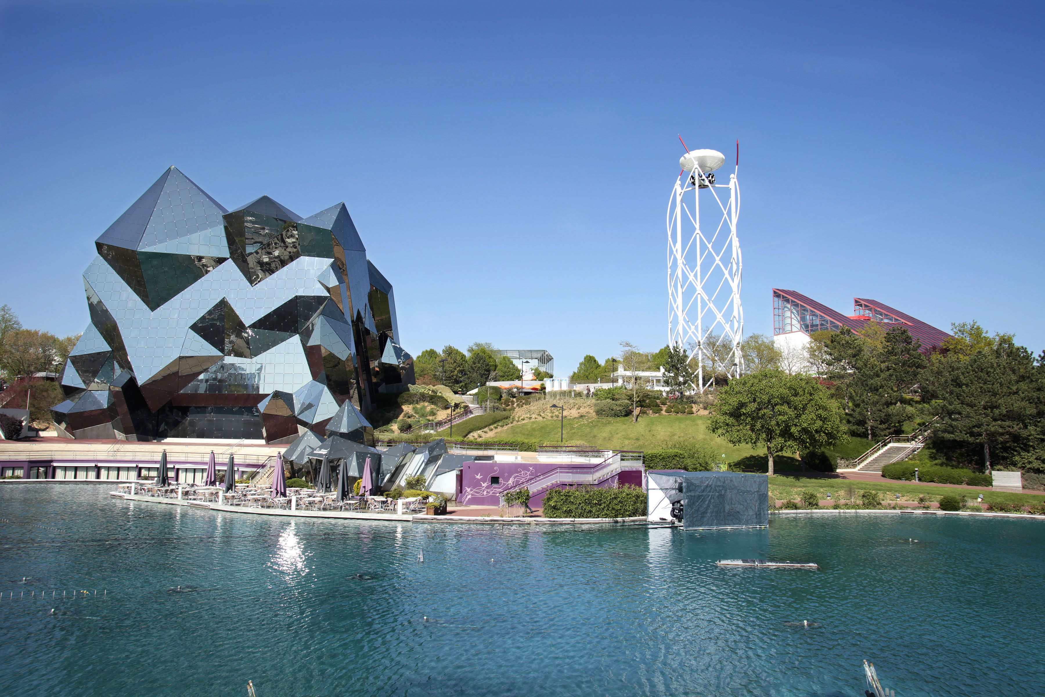 A modern building with a unique geometric facade near a body of water, a tall white tower, and a staircase leading up to a red-roofed structure.