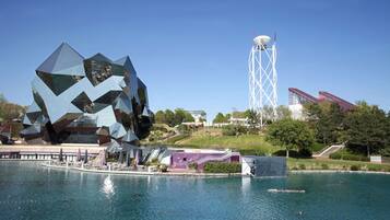A modern building with a unique geometric facade near a body of water, a tall white tower, and a staircase leading up to a red-roofed structure.